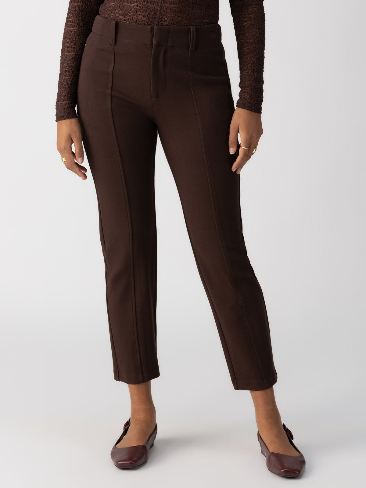 A person stands against a plain background wearing the Cassie Legging Coffee from Sanctuary Clothing, paired with a brown textured long-sleeve top and brown flat shoes. The outfit is completed with minimal jewelry.