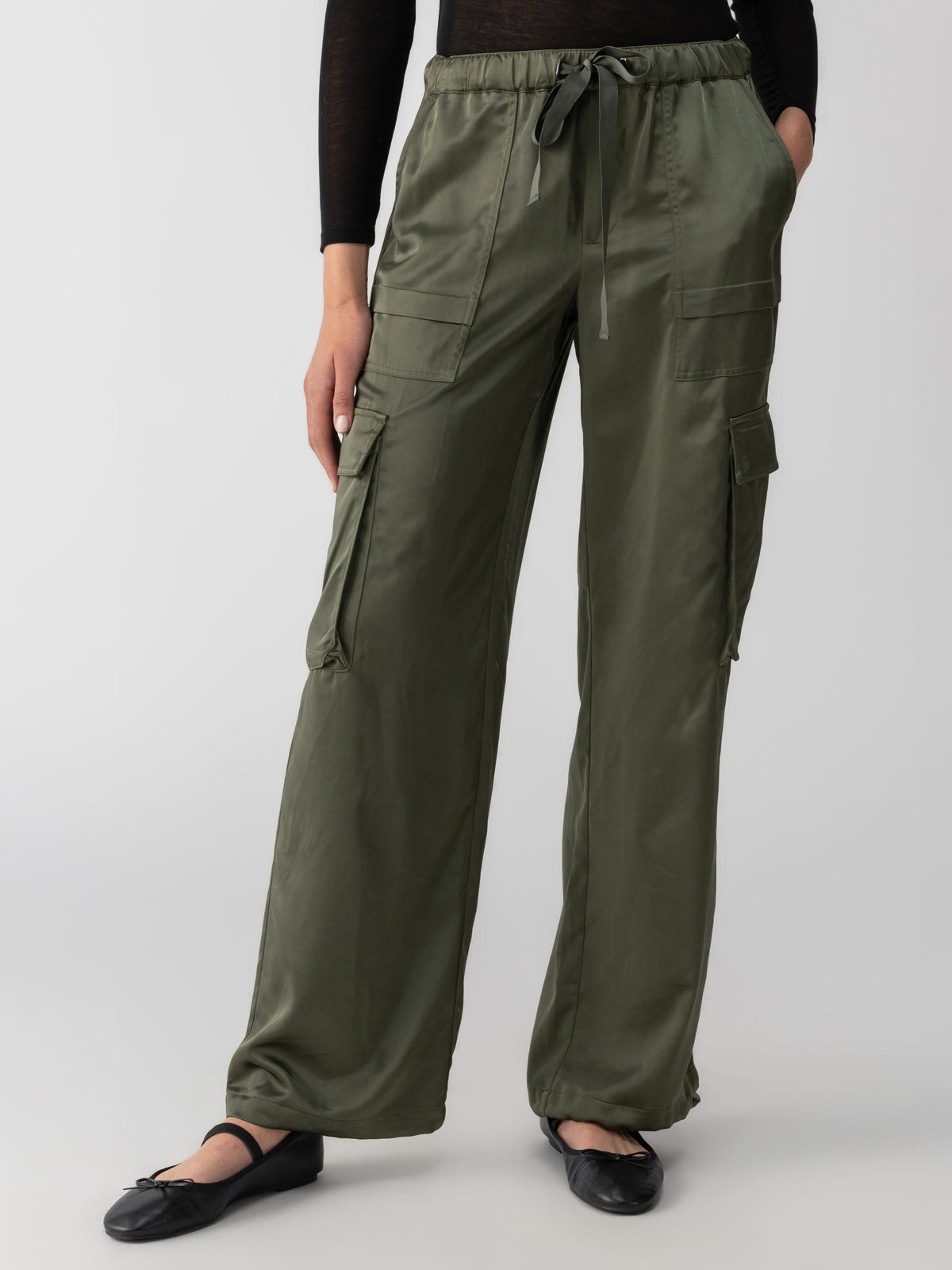 A person is wearing the "Satin Cargo Wide Leg Dark Olive" pants by Sanctuary Clothing, which feature multiple pockets and a drawstring waist. They have paired this outfit with black ballet flats and a black top. The background is plain and gray, and their left hand rests by their side.