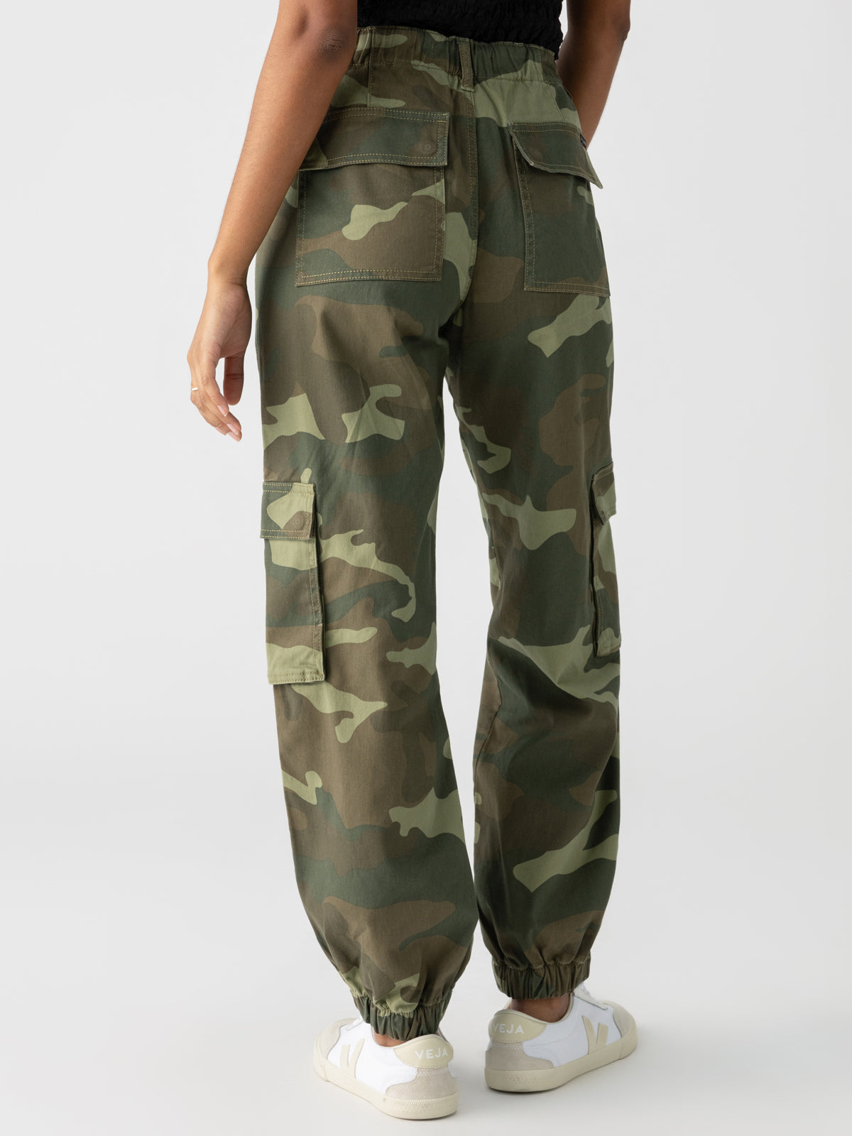 A person wearing the Cyber Cargo Renew Camo pants from Sanctuary Clothing and white sneakers is standing with their back to the camera. The pants feature multiple pockets and elastic cuffs at the ankles. The background is plain white.