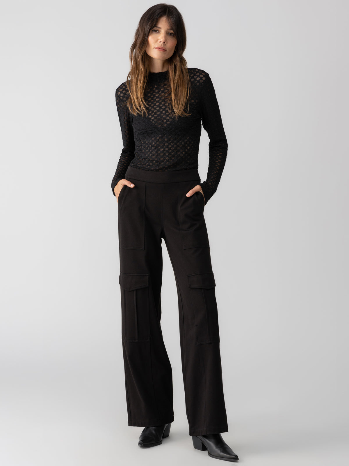 A woman with long brown hair is wearing a black, long-sleeved mesh top and the Ponte Travel Cargo Black trousers from Sanctuary Clothing. She is standing with her hands in her pockets and looking forward. The background is plain and light grey.