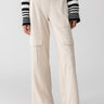 A person is modeling the Ponte Travel Cargo Toasted Almond high-waisted cargo pants from Sanctuary Clothing, featuring multiple pockets. The pants are paired with a black and white striped long-sleeve top and black flats. The background is plain and light-colored, highlighting the outfit.