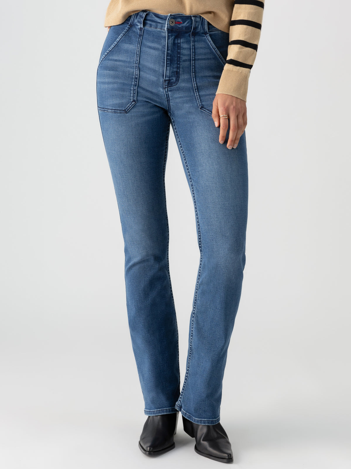 A person wears Sanctuary Clothing's Sculpted Hayden Bootcut Lucky Streak jeans, which feature front pockets and visible stitching. They pair the high-waisted blue jeans with a beige and black striped sweater, completing the look with black pointed-toe shoes. The background is light gray.