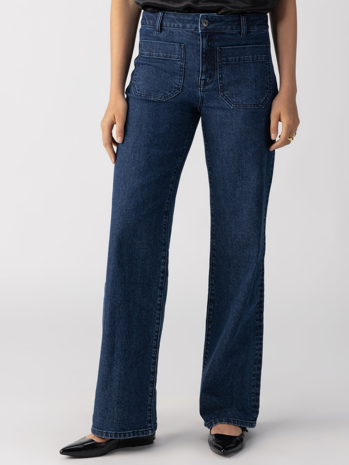 A person is shown from the waist down wearing the Marine Full Length Aspen jeans from Sanctuary Clothing, featuring a dark blue high-waisted design with a wide-leg cut. They pair these with black pointed-toe shoes and a dark top, set against a simple light gray background.