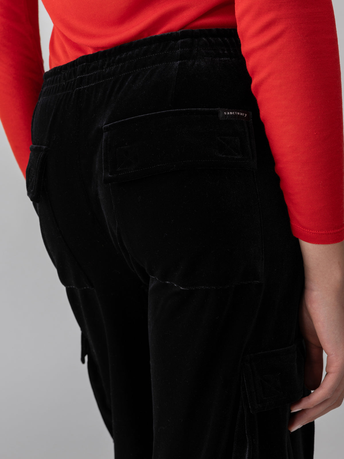 A person wearing a red long-sleeve top and Sanctuary Clothing's Relaxed Velvet Reissue Black cargo pants. The image focuses on the lower back and upper thigh area, highlighting the pockets and texture of the pants.