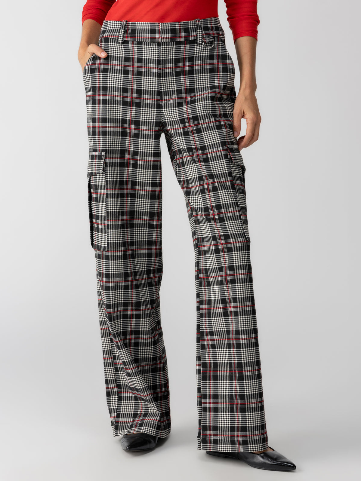 A person is wearing the Gwen Cargo Garnet Plaid trousers from Sanctuary Clothing, which feature a high-waisted, wide-leg design with a black, white, and red checkered pattern. The pants have side cargo pockets. The wearer is also dressed in a long-sleeve red top and black pointed-toe shoes.