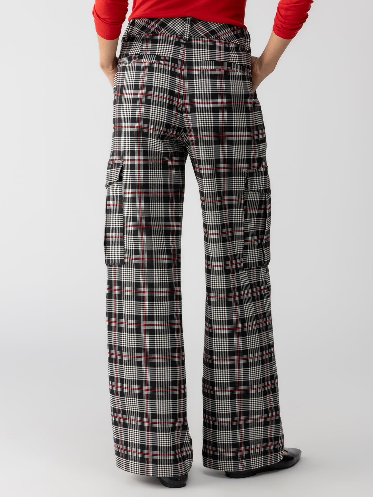 A person wearing the Gwen Cargo Garnet Plaid pants from Sanctuary Clothing, featuring a high waist, wide legs, and multiple pockets including cargo pockets on each side, stands with their back to the camera. They are also wearing a red long-sleeved top. The background is plain and light-colored.