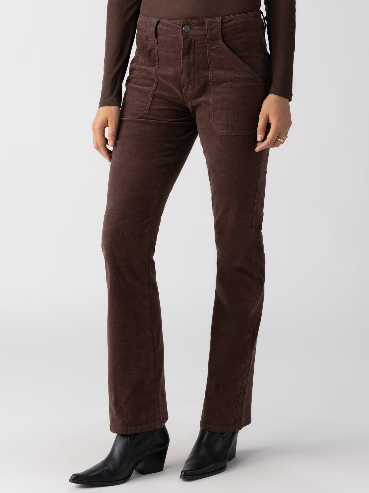 A person is wearing the Corduroy Hayden Bootcut Brown Sugar pants by Sanctuary Clothing, featuring a high-waisted design and front pockets. They are also donning a dark long-sleeve shirt and black ankle boots. The background is plain and light-colored, with only the person's lower torso and legs visible.