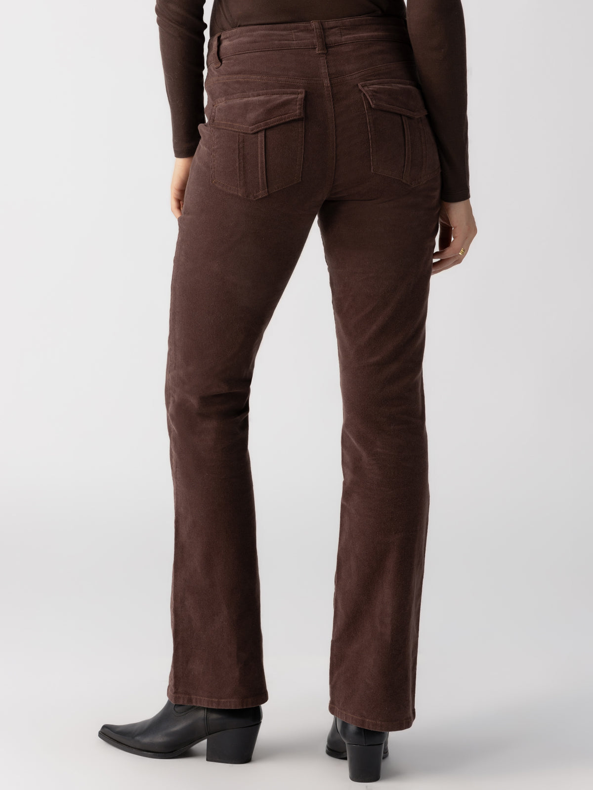 A person is shown from the back wearing Sanctuary Clothing's Corduroy Hayden Bootcut pants in Brown Sugar, paired with a dark long-sleeve top and black ankle boots. The corduroy pants feature visible back pockets, and the individual is standing against a neutral background.