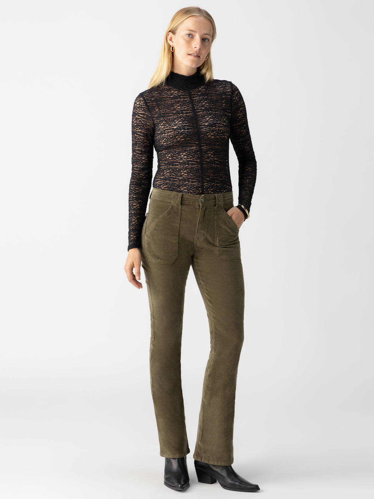 A person with long blonde hair is dressed in a black, sheer lace, long-sleeved top paired with the Sanctuary Clothing Corduroy Hayden Bootcut pants in Burnt Olive, featuring visible pockets. They complete their look with black ankle boots. The background is a plain light grey.
