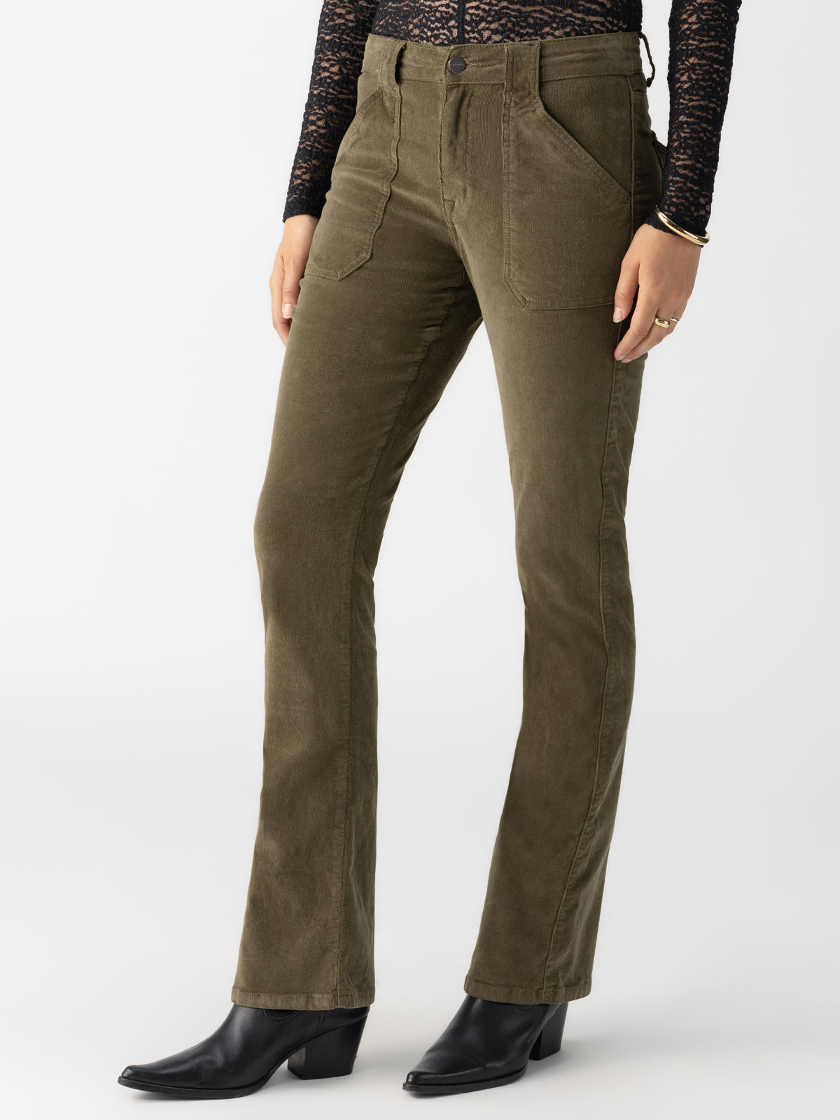 A person wears the Corduroy Hayden Bootcut Burnt Olive from Sanctuary Clothing, paired with a black lace long-sleeve top and black heeled boots, against a plain white background.