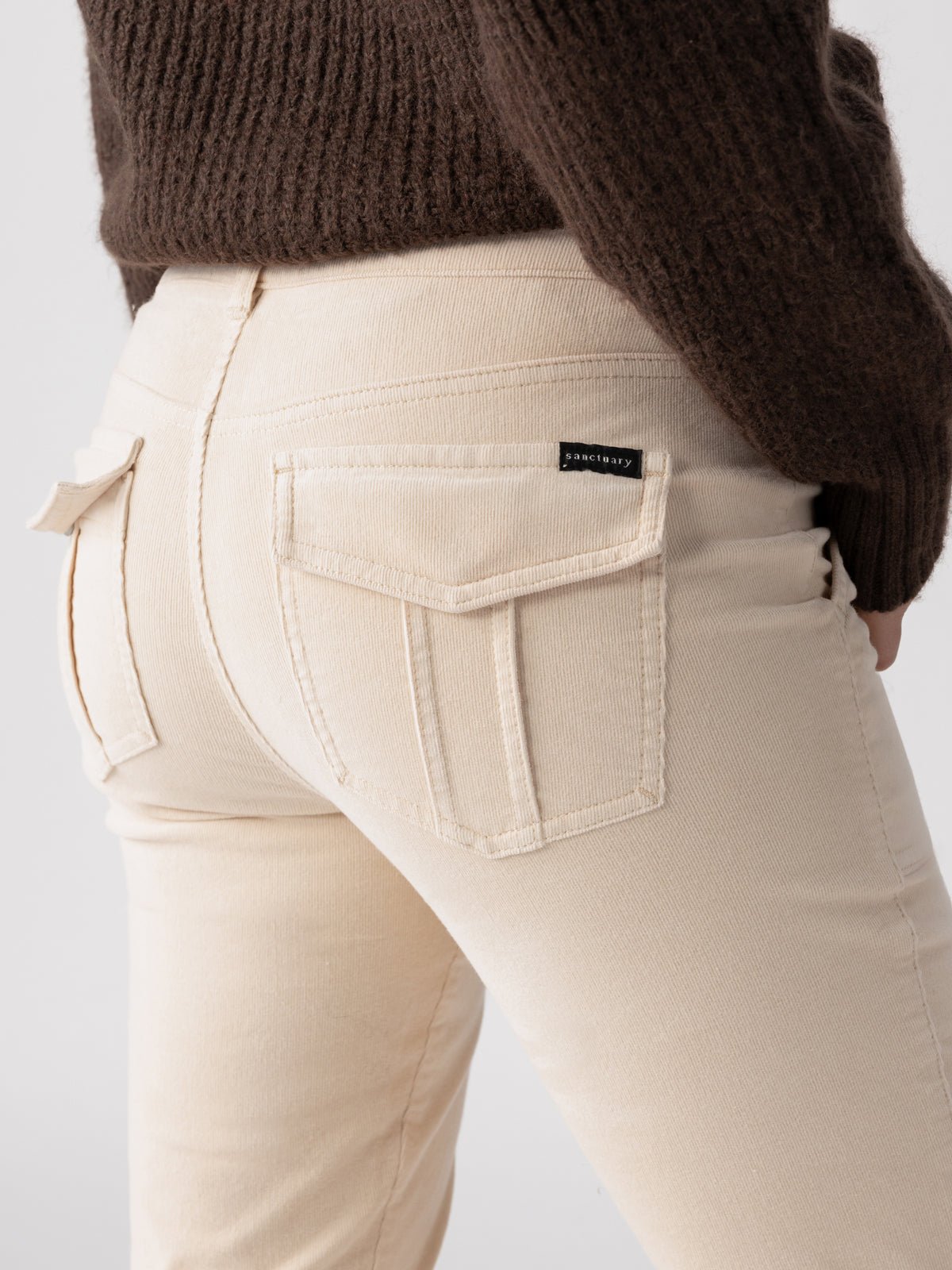 A person wearing Sanctuary Clothing's Corduroy Hayden Bootcut Toasted Almond pants with visible back pockets and a black label. They are also wearing a dark brown sweater. The photo is focused on the lower back and upper thigh area.