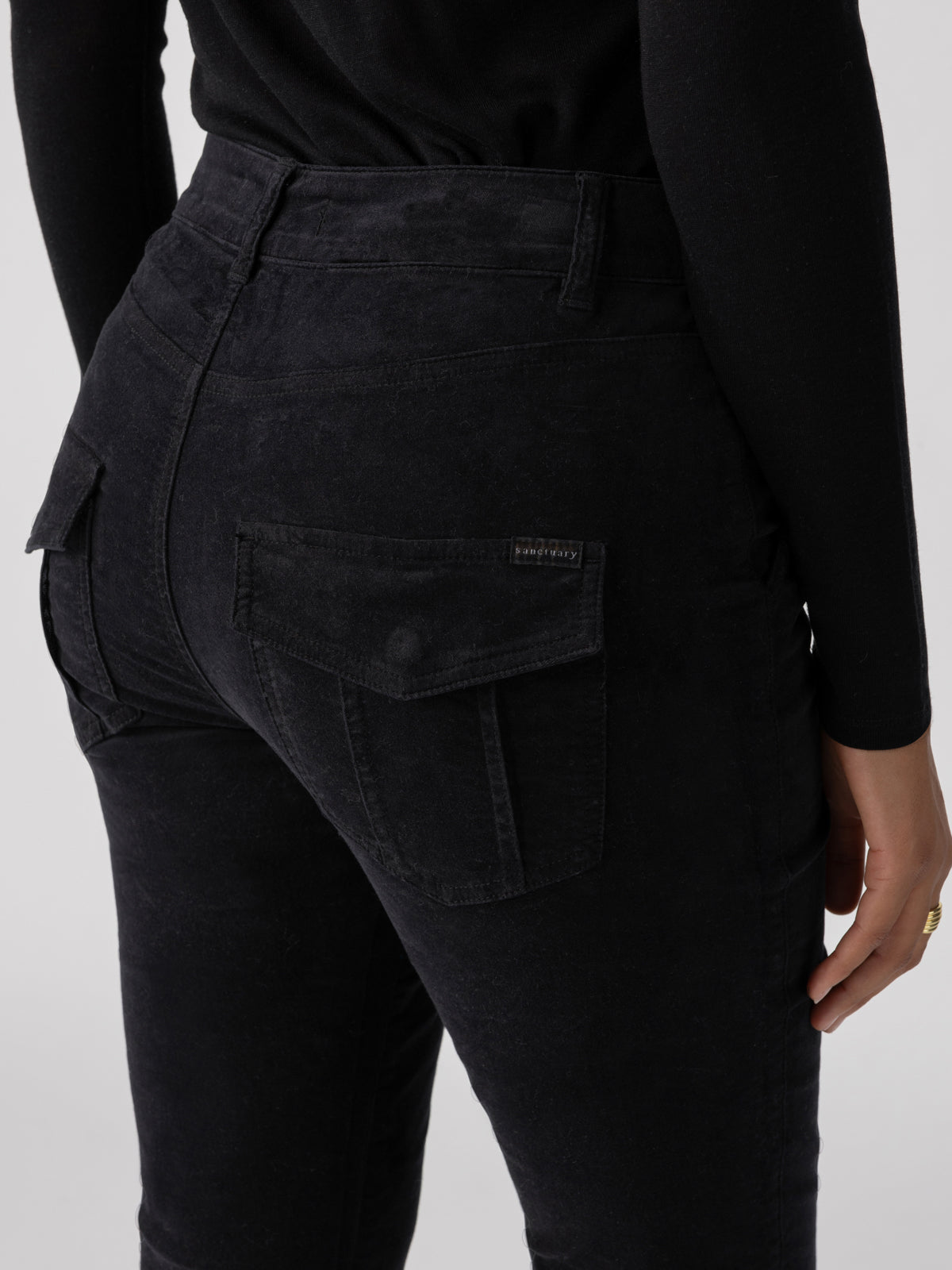 A person is standing, facing away, wearing black Velveteen Hayden Bootcut Pants from Sanctuary Clothing, made of a textured fabric. The pants have flap pockets on the back, and the person is also wearing a black long-sleeve top.