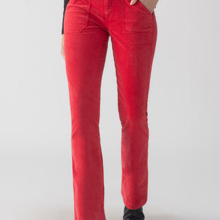 A person wearing the Velveteen Hayden Bootcut Pant in Mars Red by Sanctuary Clothing paired with a black top, standing against a plain white background. The high-waisted, flared red corduroy pants feature a button and zipper closure along with front patch pockets, and the person is posing with one hand in their pocket.