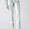 An individual dressed in the Sanctuary Clothing Metallic Hayden Bootcut Pant in Silver paired with a textured gray sweater and black heeled boots stands against a plain background.