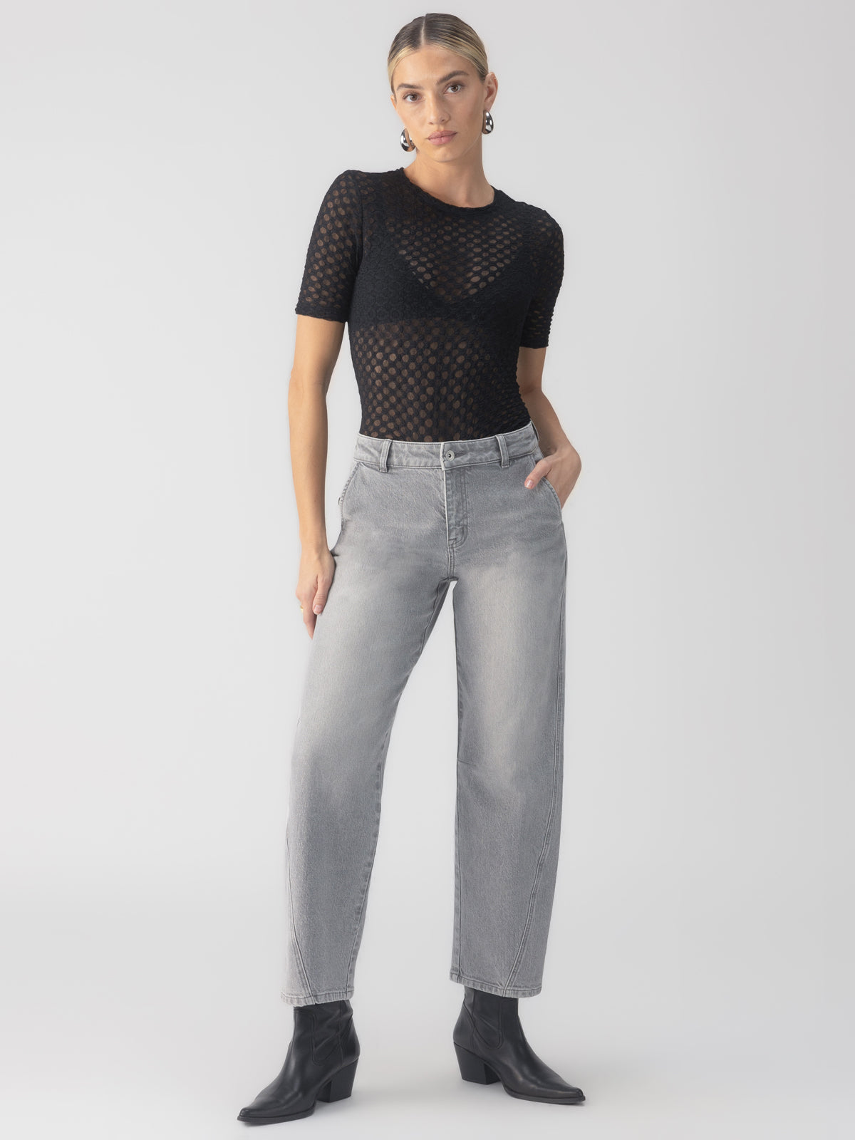 A person stands confidently wearing a black mesh short-sleeve top, Sanctuary Clothing's twisted seam jeans barrel in grey mist, and black ankle boots. Their left hand rests in their pocket, and they have short, tied-back hair. The background is plain white.