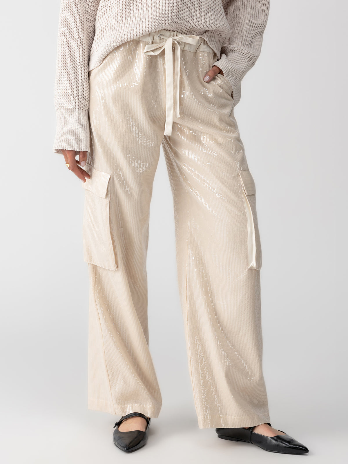 A person wearing the Pull On Sequin Cargo Pant in Frosted Almond by Sanctuary Clothing, featuring high-waisted design and tie belt, is styled with a loose-knit sweater and black pointed flats. The outfit is showcased against a plain background.