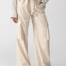 A person wearing the Pull On Sequin Cargo Pant in Frosted Almond by Sanctuary Clothing, featuring high-waisted design and tie belt, is styled with a loose-knit sweater and black pointed flats. The outfit is showcased against a plain background.