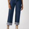 A person wearing the Luna Cuffed Denim Pant Solstice from Sanctuary Clothing, a pair of high-waisted blue jeans with cuffed hems and button details, is paired with an untucked blue shirt and beige ankle boots against a plain white background.