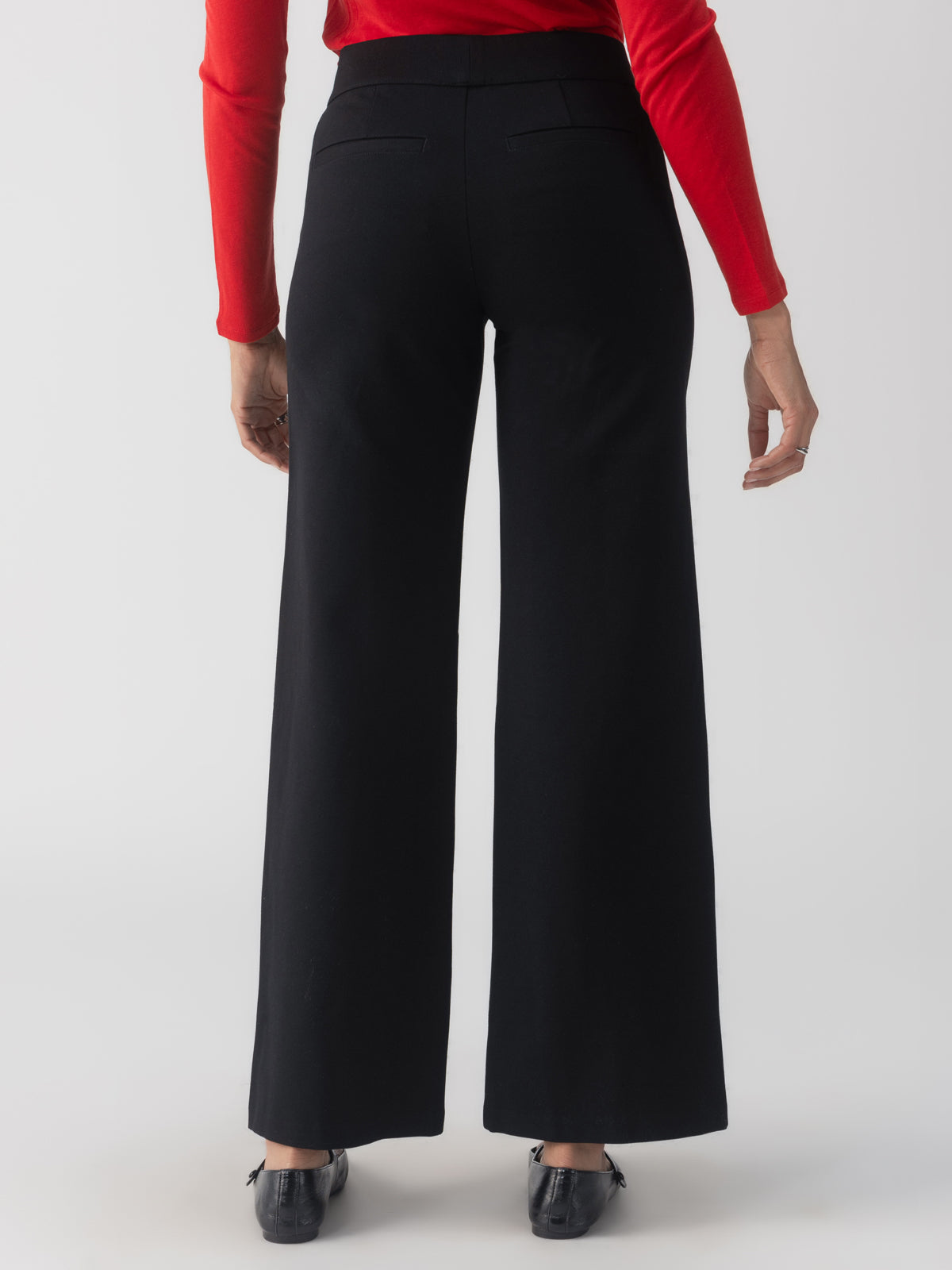 A person wearing a red long-sleeve top and the Runway Ponte Trouser Black from Sanctuary Clothing is seen from behind on a light gray background, paired with black shoes.