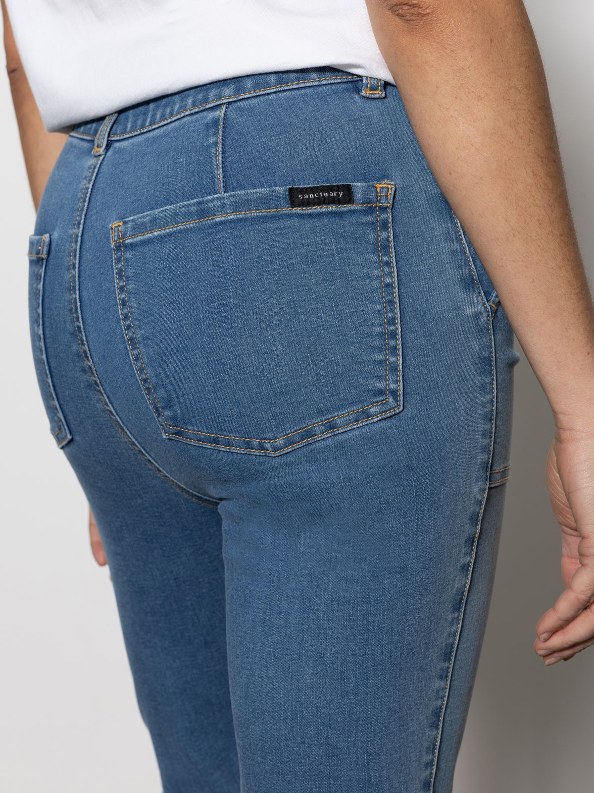 A close-up shot features someone wearing the "sculpted hayden straight crop best in show" jeans by Sanctuary Clothing and a white top. The focus is on the back pocket area, adorned with a small black label, as the individual stands against a neutral background.