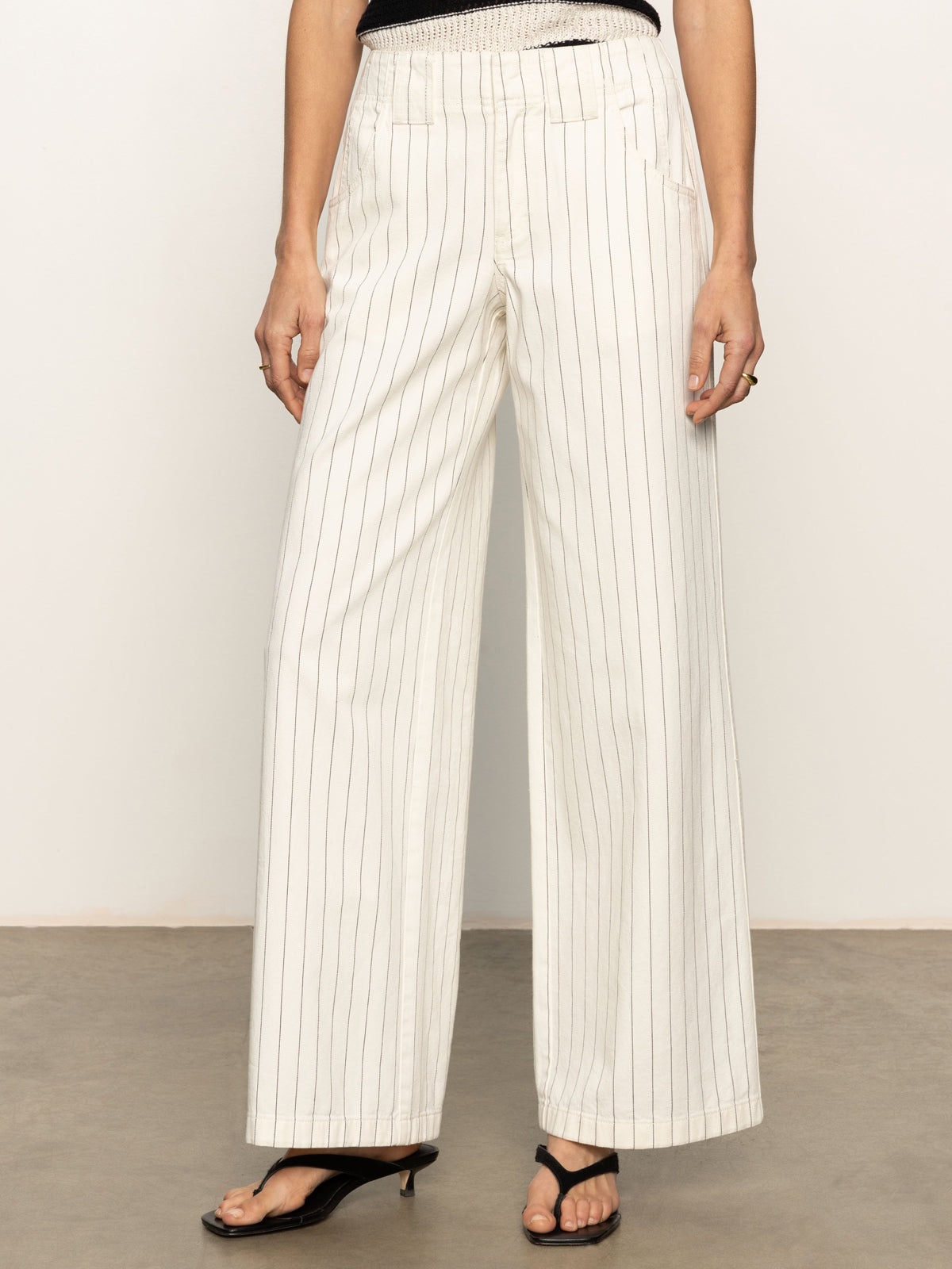 An individual is dressed in arden wide leg jeans by Sanctuary Clothing, featuring bleached white pinstripes. The pants are complemented by black heeled sandals against a plain light-colored wall and floor.