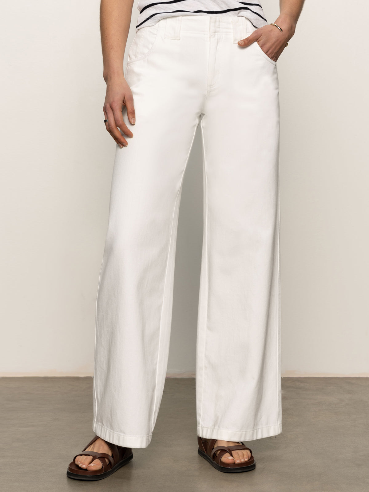 Person wearing Sanctuary Clothing's arden wide leg jeans in brilliant white, with one hand in pocket, standing on a concrete floor. They're also wearing a striped top and black sandals. The image is cropped above the shoulder.