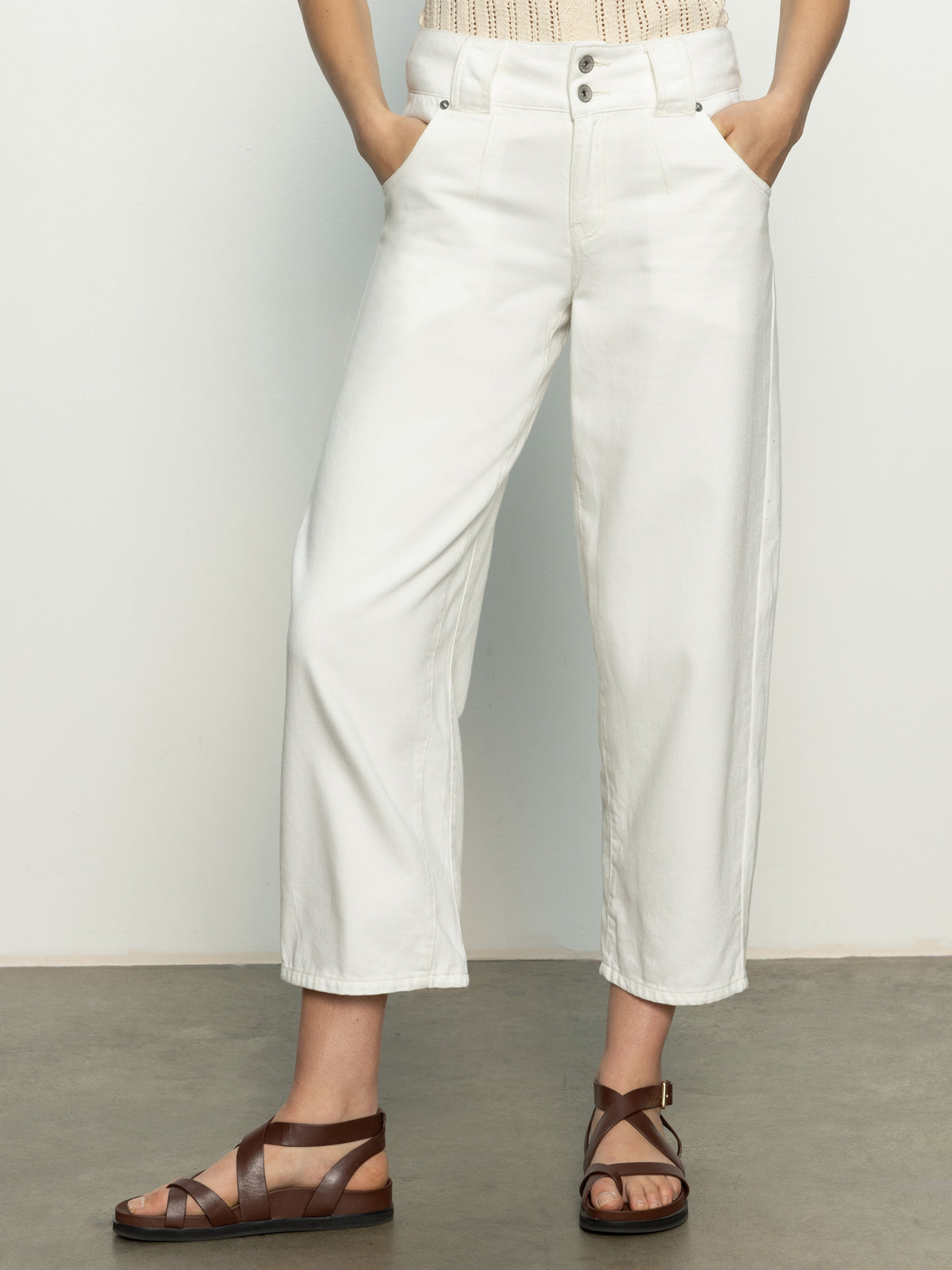 A person wears Sanctuary Clothing's side car barrel jeans in bleached white, pockets filled, styled with brown strappy sandals. The photo highlights the lower half of the outfit against a plain, light background and is cropped at the shoulders.