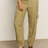 A person in the Sanctuary Clothing's "take over linen jogger" pants in olive green, featuring side pockets and elastic cuffs, is wearing brown sandals on a gray floor. Their hand rests casually on their thigh while donning a white shirt.