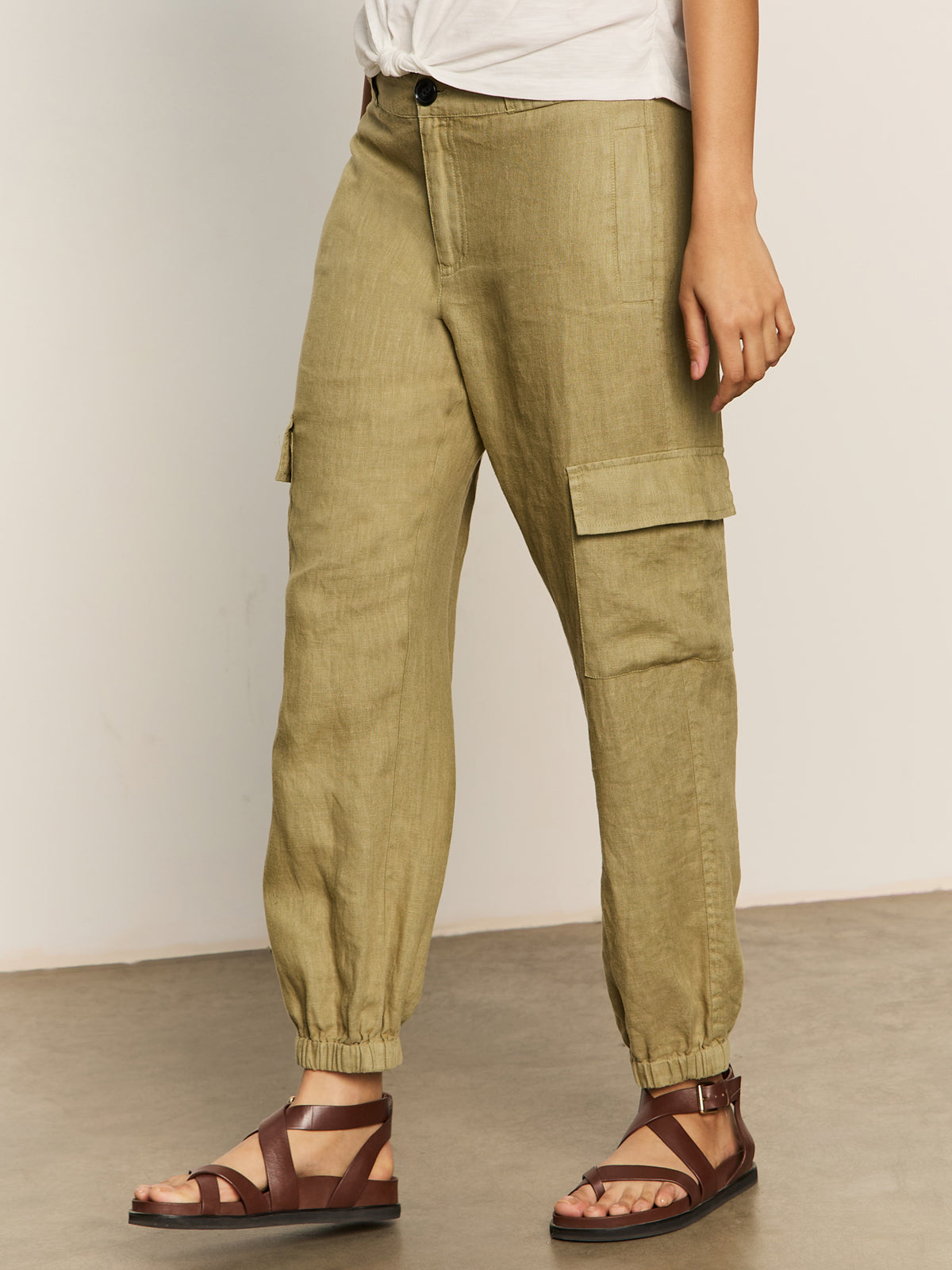A person in Sanctuary Clothing's "take over linen jogger standard rise pant" in bamboo is wearing brown sandals. The pants feature large side pockets and elastic cuffs. They're standing on a beige floor with a white wall background.