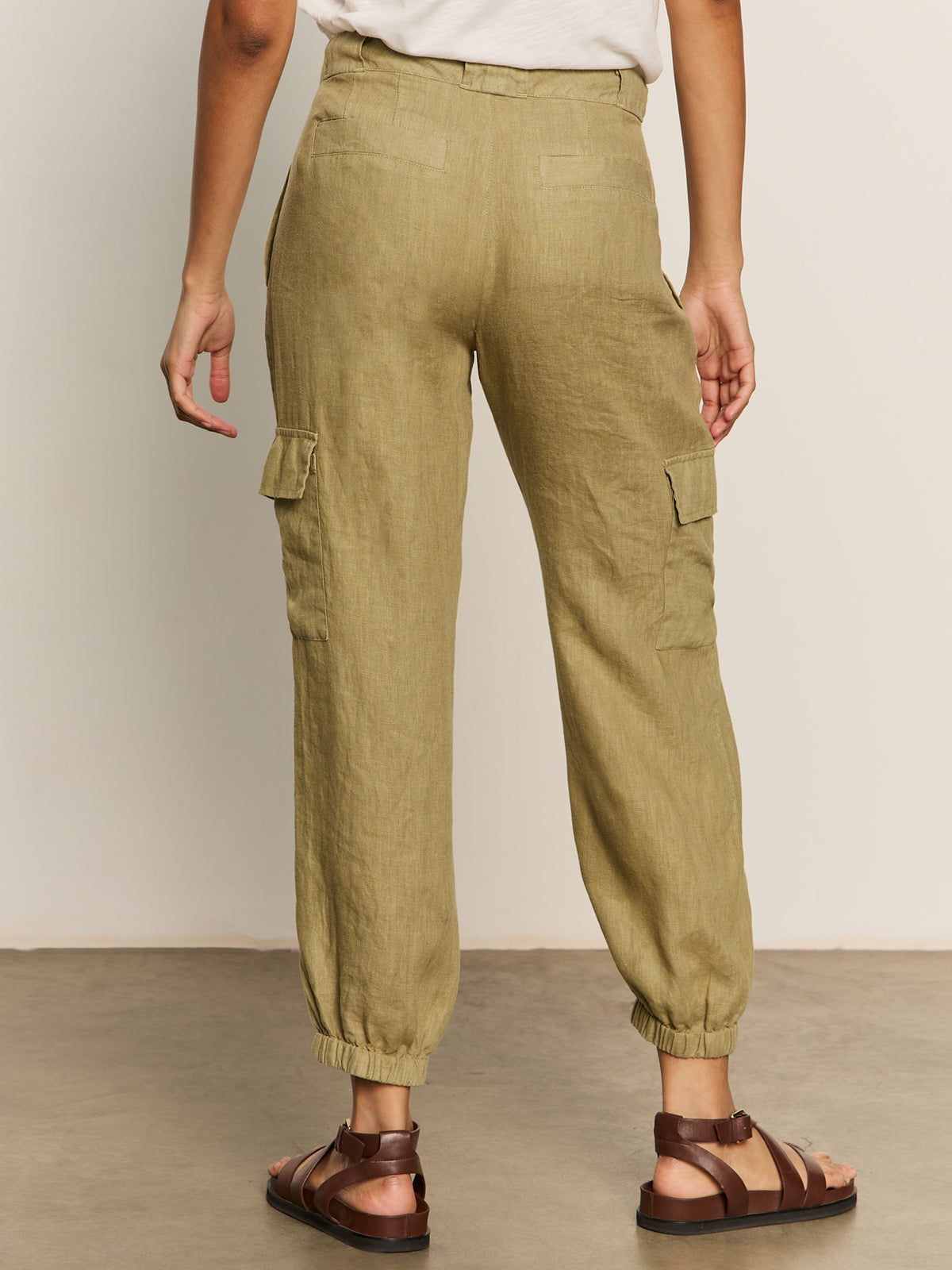 A person is seen from the rear in Sanctuary Clothing's "Take Over" linen jogger pants in bamboo, paired with a casual white top and brown sandals, against a neutral background.