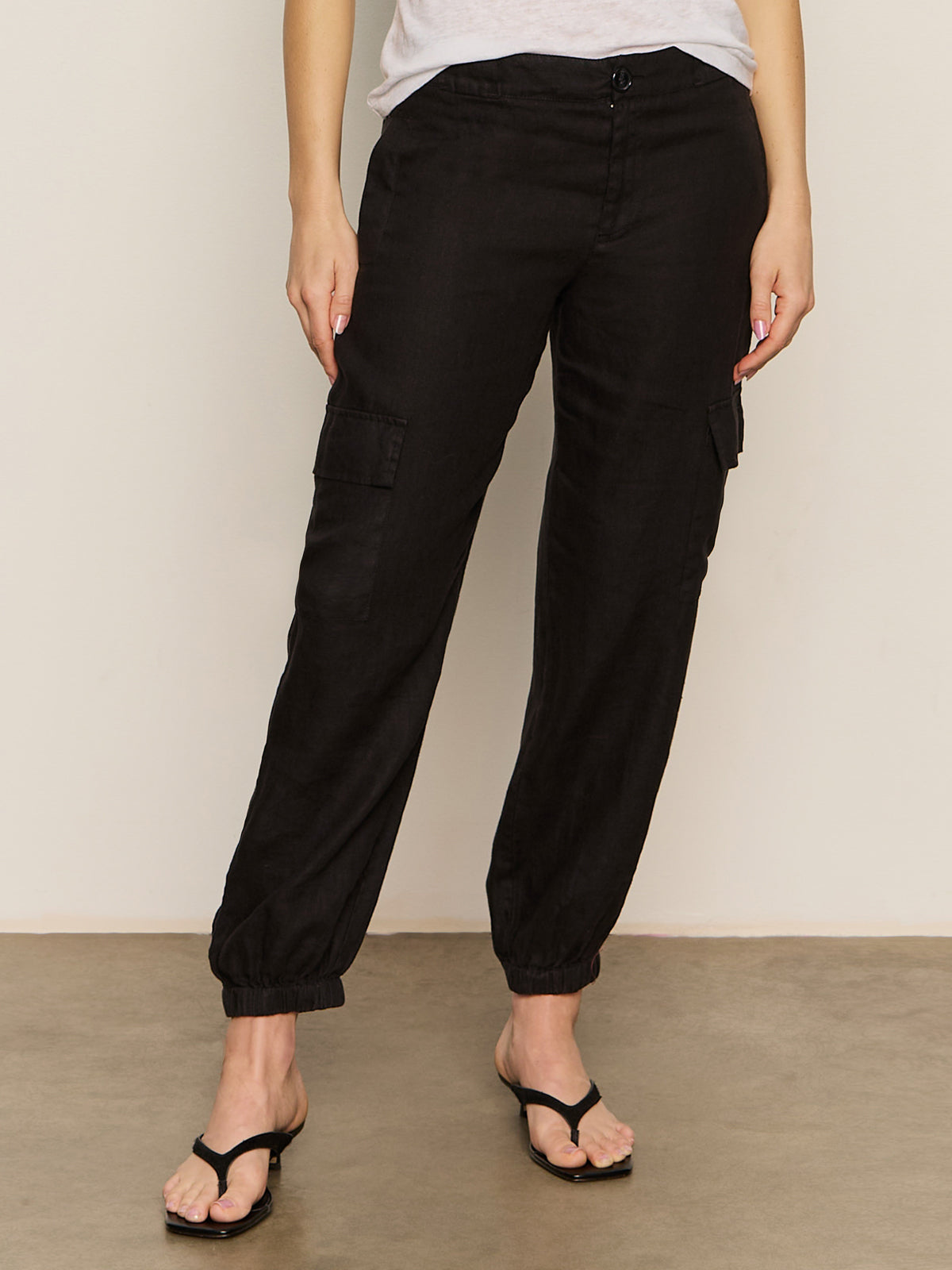 A person wearing the black take over linen jogger standard rise pant from Sanctuary Clothing and black sandals stands against a neutral background. Their hands hang casually by their sides. The pants feature multiple pockets, while the sandals boast a simple thong design.