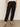 A person wearing the black take over linen jogger standard rise pant from Sanctuary Clothing and black sandals stands against a neutral background. Their hands hang casually by their sides. The pants feature multiple pockets, while the sandals boast a simple thong design.