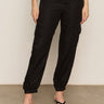 A person wearing the black take over linen jogger standard rise pant from Sanctuary Clothing and black sandals stands against a neutral background. Their hands hang casually by their sides. The pants feature multiple pockets, while the sandals boast a simple thong design.