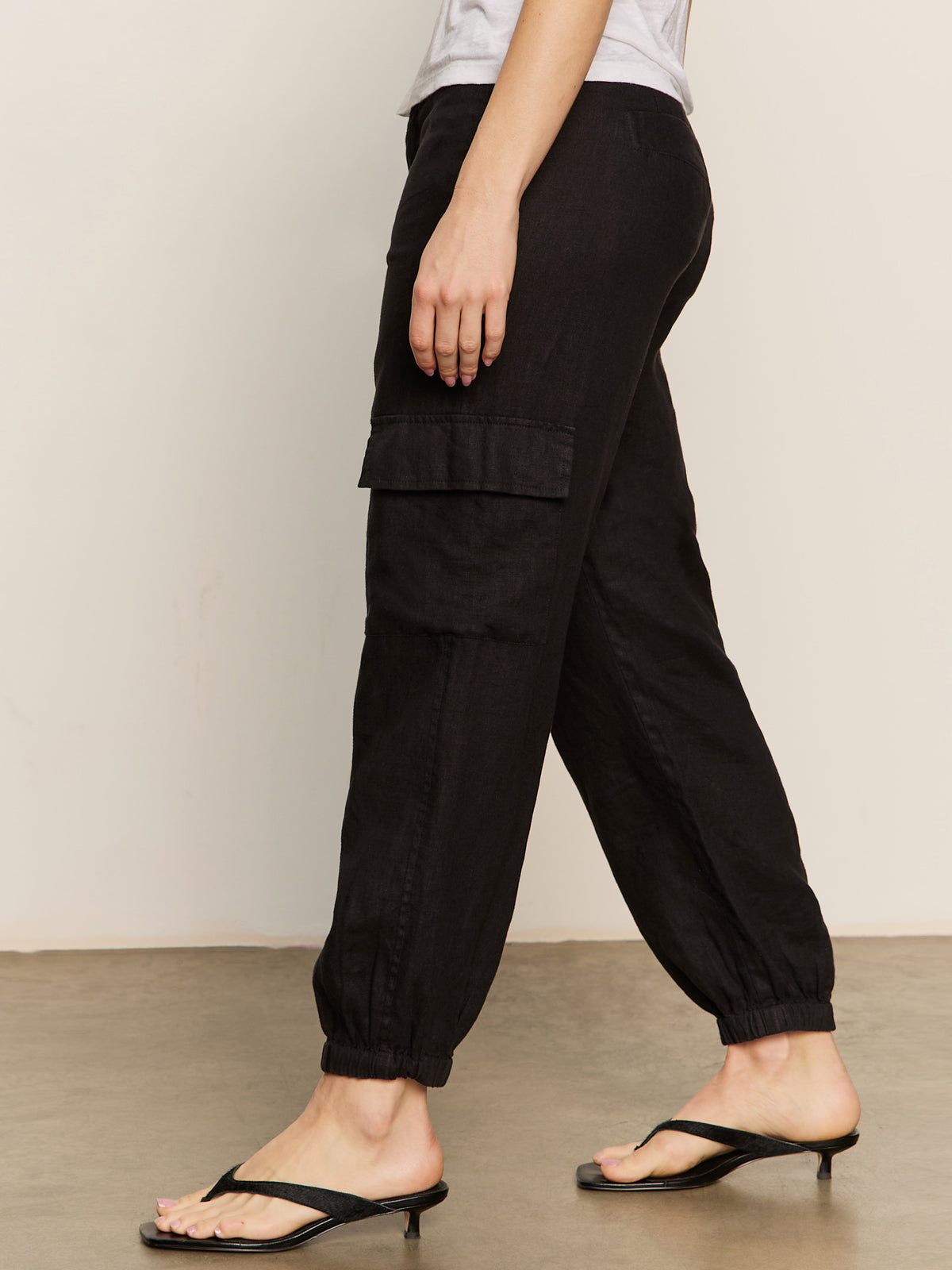 A person wearing Sanctuary Clothing's black "take over linen jogger standard rise pant" and black heeled sandals walks on a concrete floor, partially visible from the waist down, paired with a light-colored top.