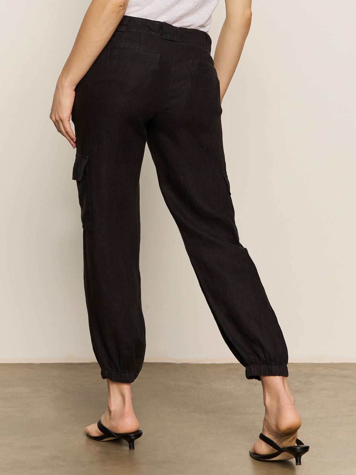 A person wearing Sanctuary Clothing's "Take Over Linen Jogger" in black, paired with black heels, is shown from the back. The joggers feature side pockets and elastic cuffs. They stand on a beige floor with a light neutral background.