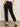A person wearing Sanctuary Clothing's "Take Over Linen Jogger" in black, paired with black heels, is shown from the back. The joggers feature side pockets and elastic cuffs. They stand on a beige floor with a light neutral background.