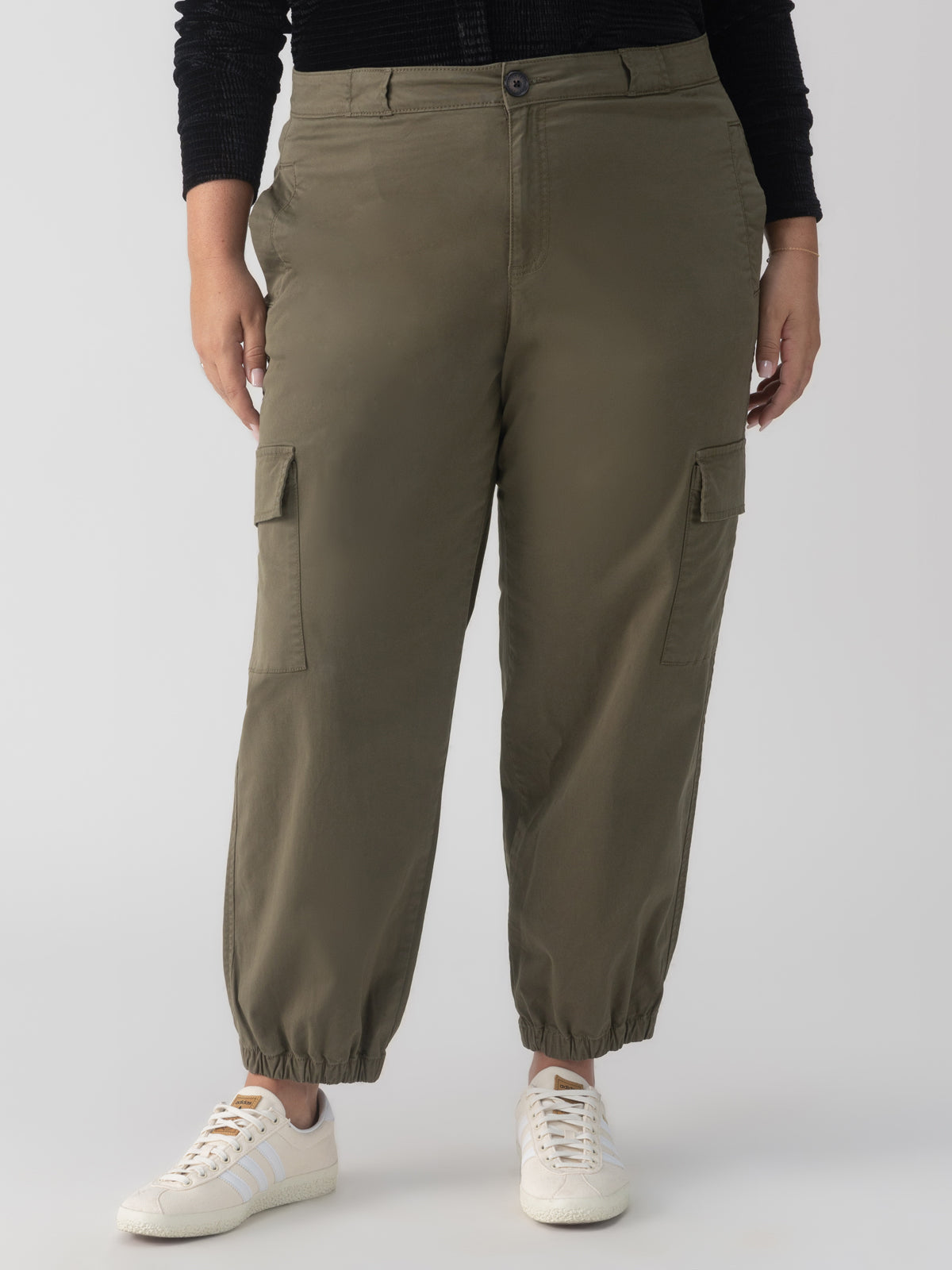 A person is shown wearing the Take Over Jogger Kalamata from the Sanctuary Clothing Inclusive Collection, featuring elasticized cuffs and side pockets. They are also sporting a black top and white sneakers. The background is a plain, light gray color.