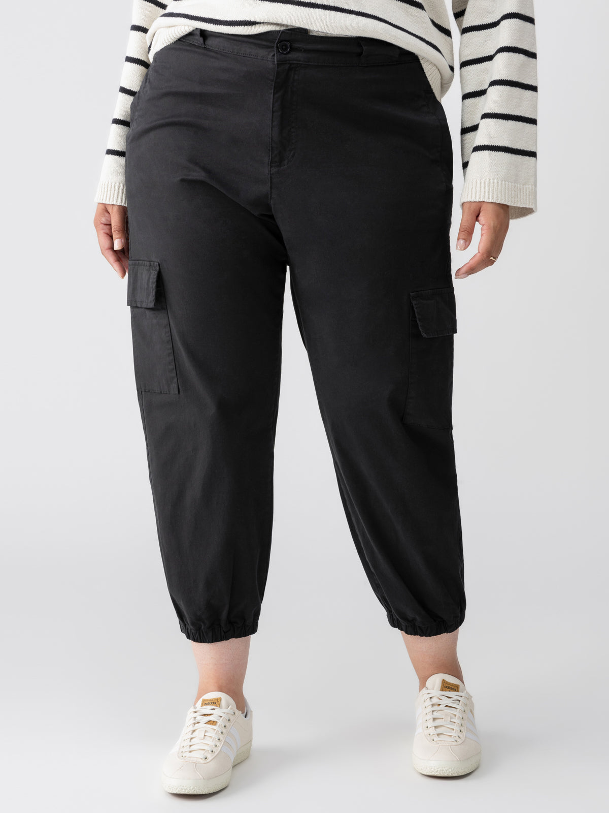 A person is wearing a white sweater with black horizontal stripes, the Take Over Jogger Black Inclusive Collection by Sanctuary Clothing, and white sneakers. The joggers have side pockets and are cropped at the ankle. The background is plain white.