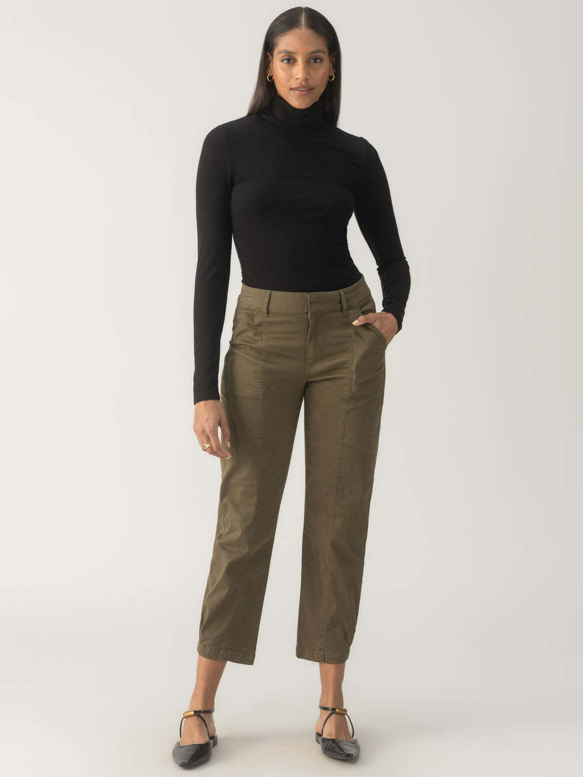 A person stands against a plain background, wearing a black turtleneck, the Sahara Tapered Pant in Burnt Olive by Sanctuary Clothing, and black shoes. They have long hair and are posing with one hand in their pocket, looking confidently at the camera.