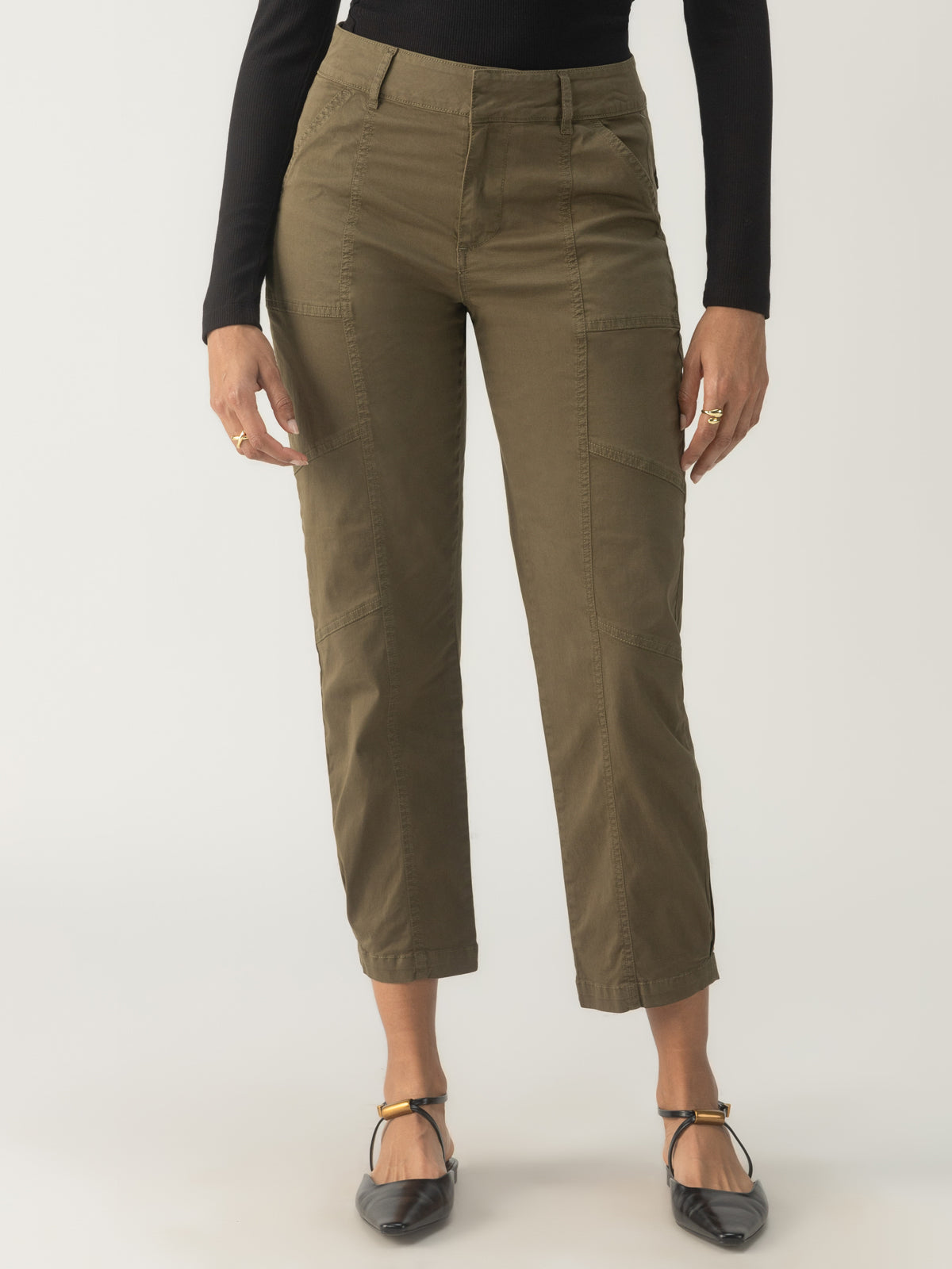 A person is standing in the Sahara Tapered Pant in Burnt Olive by Sanctuary Clothing, alongside a black long-sleeve top. These pants feature multiple pockets, and the individual complements their look with black flat shoes featuring ankle straps.