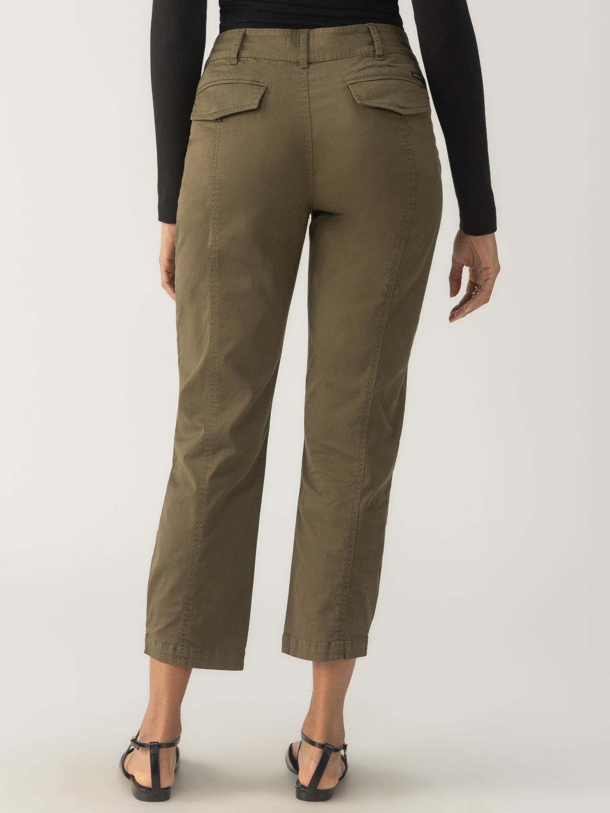 A person is wearing the Sahara Tapered Pant in Burnt Olive from Sanctuary Clothing, paired with a black long-sleeve top, and two back pockets are visible. They are standing against a neutral background, complemented by black sandals.