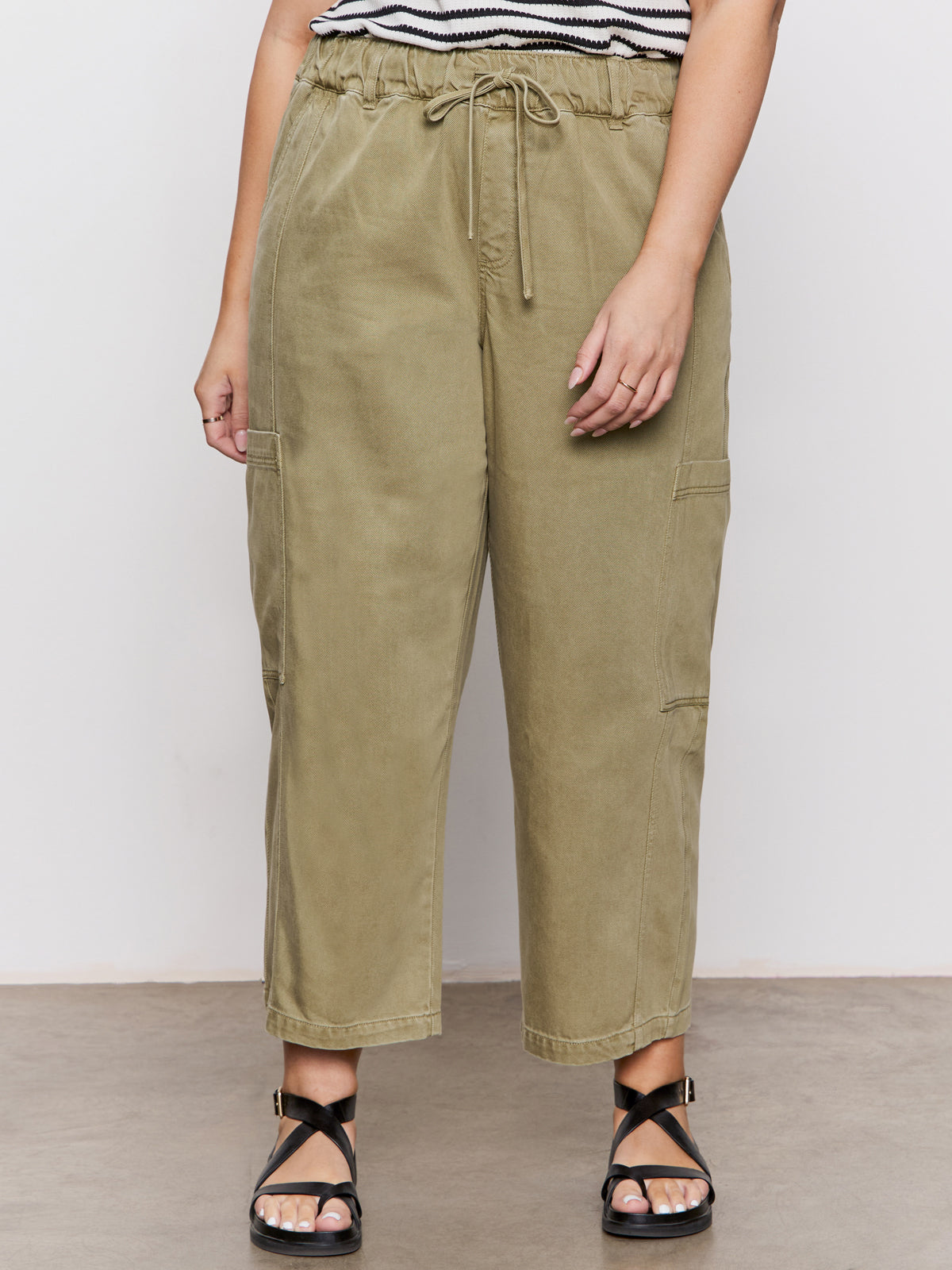 A person wearing Sanctuary Clothing’s area standard rise pant bamboo extended sizing in olive, paired with black strappy sandals, stands against a plain light background. Only the lower half of their body is visible.
