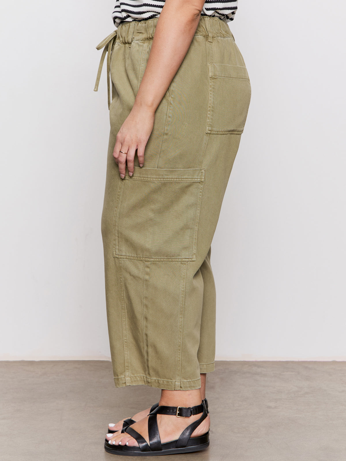 A person stands sideways in Sanctuary Clothing's area standard rise pant bamboo extended sizing, featuring olive green cargo style, paired with a black and white striped top and black sandals on a concrete floor against a plain light background.