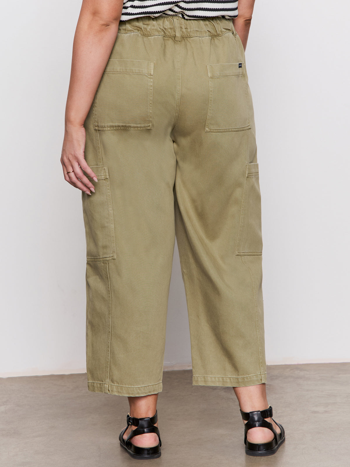 Someone is wearing the Sanctuary Clothing area standard rise pant bamboo extended sizing in olive green, with large back and side pockets, paired with black sandals and a striped top, standing against a plain light background.