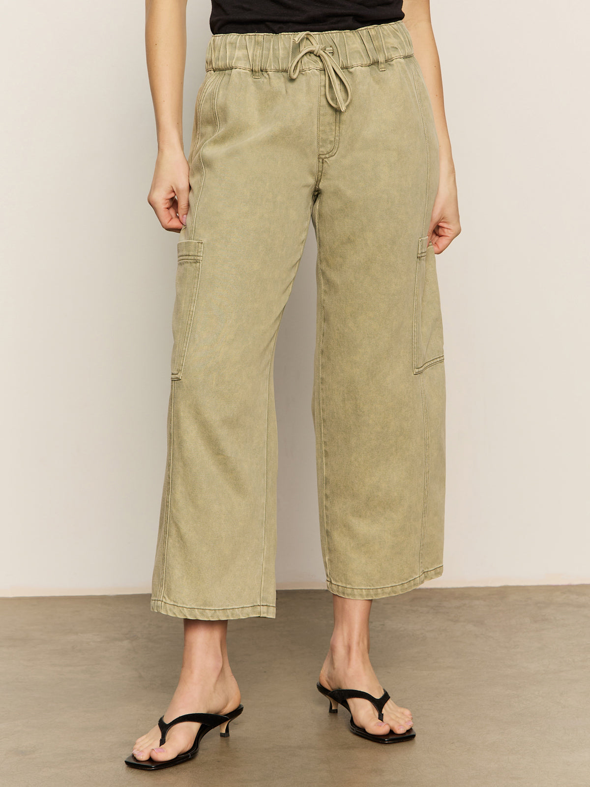 A person wears Sanctuary Clothing's area standard rise pant bamboo in light olive green with a drawstring waist and wide legs, paired with black heeled sandals, standing on a beige floor against a plain light wall.