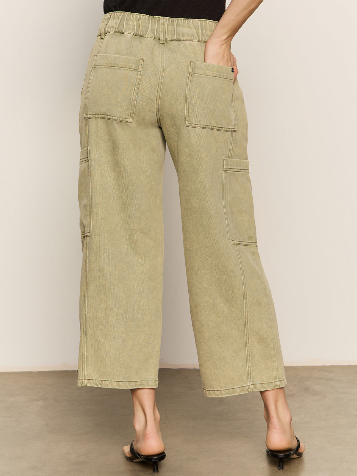 A person wearing Sanctuary Clothing's area standard rise pant bamboo—high-waisted, wide-leg light olive cargo pants with large back and side pockets—paired with black heeled sandals and one hand in a pocket.
