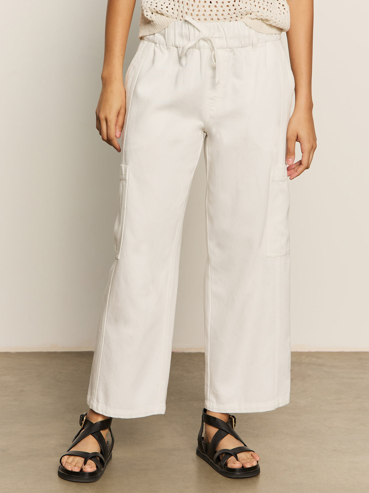 The lower half of a person is shown wearing Sanctuary Clothing's area standard rise pant in bleached white, featuring a drawstring waist, side pockets, and paired with black strappy sandals.