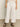 The lower half of a person is shown wearing Sanctuary Clothing's area standard rise pant in bleached white, featuring a drawstring waist, side pockets, and paired with black strappy sandals.