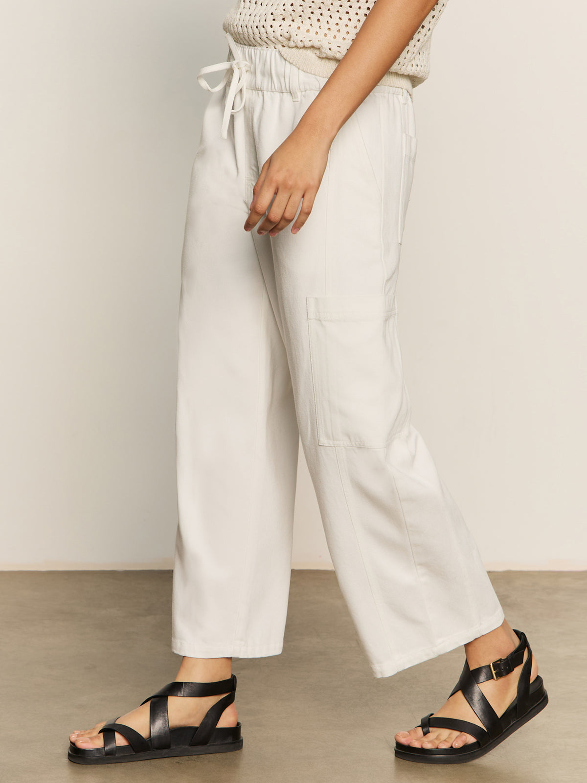A person wears Sanctuary Clothing's area standard rise pant in bleached white, styled with black strappy sandals and a sleeveless beige dotted top. Only the lower half of their body is visible as they stand on a smooth floor.