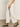 A person wears Sanctuary Clothing's area standard rise pant in bleached white, styled with black strappy sandals and a sleeveless beige dotted top. Only the lower half of their body is visible as they stand on a smooth floor.